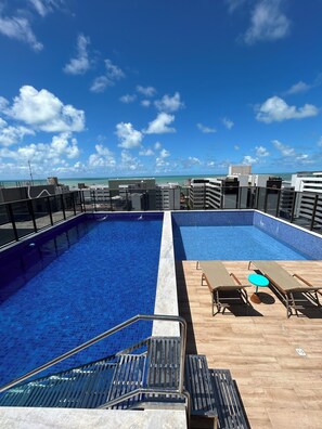 A rooftop pool with two separate sections, a wooden deck with lounge chairs and a small table, and a clear view of the ocean and buildings in the background. - Melhores Flats - Agradável perto do mar (João Pessoa)