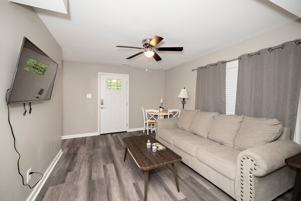 A living room with a beige sofa, a wooden coffee table, a ceiling fan, and a television mounted on the wall.
