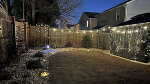 Un jardin avec une clôture en bois, un chemin de gravier et des guirlandes lumineuses.