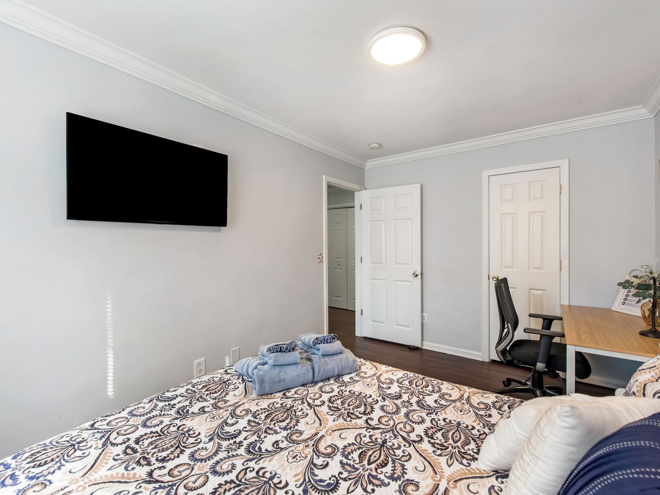 Ein Schlafzimmer mit einem Bett, einem Schreibtisch und einem Stuhl. Es gibt zwei Türen und einen an der Wand befestigten Fernseher.
