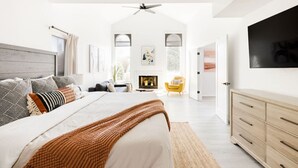 Une chambre spacieuse avec un grand lit, une tête de lit en bois, une cheminée, un fauteuil jaune et un téléviseur fixé au mur.