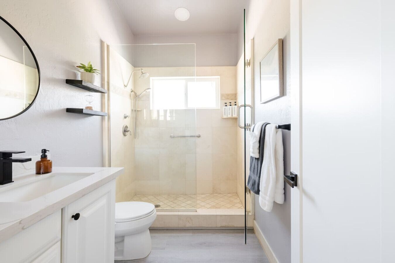 A modern bathroom with a white vanity, a round mirror, a toilet, a glass-enclosed shower, and a towel rack.