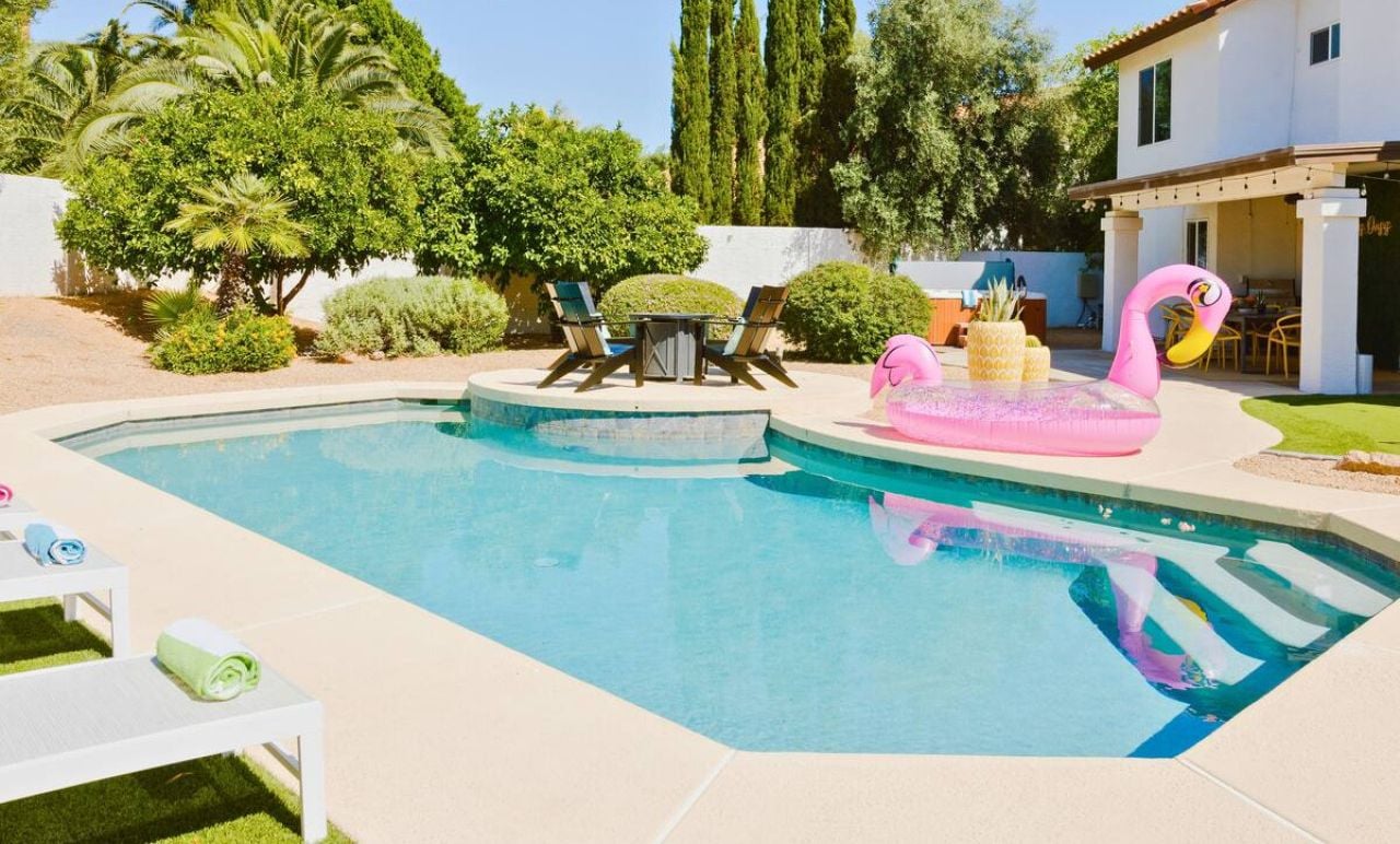 Ein Swimmingpool mit Liegestühlen und einem Flamingo-Schwimmpfad, umgeben von viel Grün und einem weißen Gebäude.
