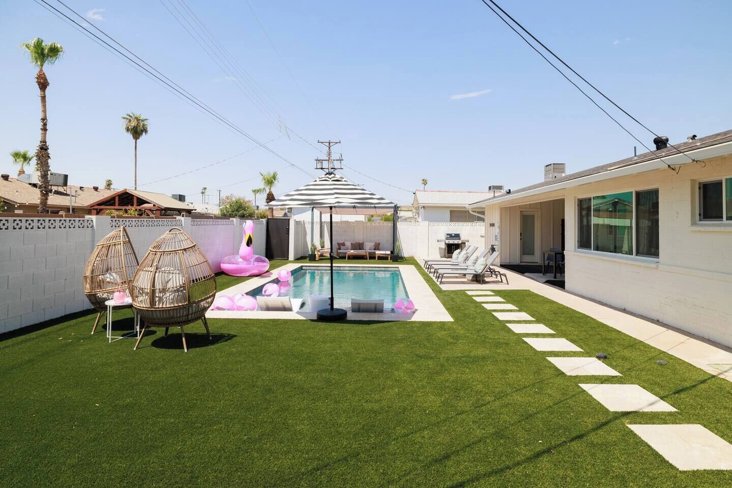 A backyard with a swimming pool, artificial grass, lounge chairs, and a pergola.