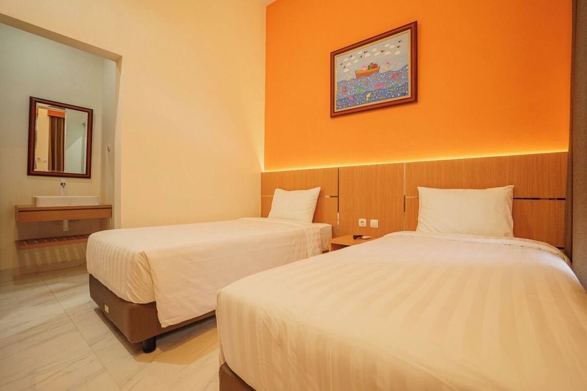 Ein Hotelzimmer mit zwei Betten, einem Badezimmer mit Spiegel und einer orangefarbenen Wand mit einem gerahmten Bild.