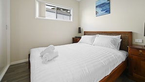 A neatly made bed with white linens and rolled towels, a wooden headboard, a nightstand with a lamp, and a painting of a wave on the wall. - Cabin 60 - Lake Conjola (Lake Conjola)