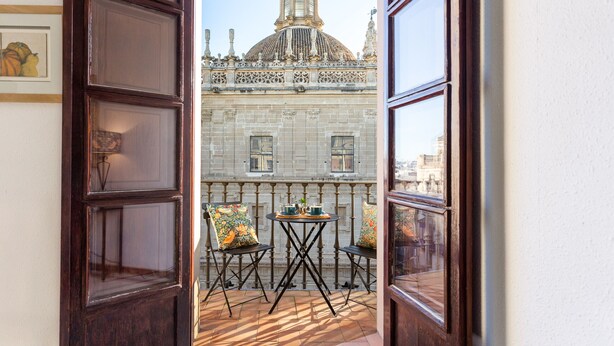 Une porte ouverte donnant sur un balcon avec une table et des chaises, une vue sur un bâtiment historique doté d’un dôme et d’un clocher, et un tableau encadré accroché au mur.