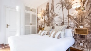 Une chambre avec un grand lit, une fresque murale représentant une végétation tropicale, une table de chevet avec une lampe et une porte dotée d’une poignée.