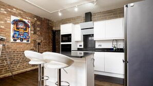 Una cucina moderna con mobili bianchi, un piano di lavoro nero e un frigorifero in acciaio inox. Una pittura adorna la parete in mattoni ed un taburete è in primo piano.