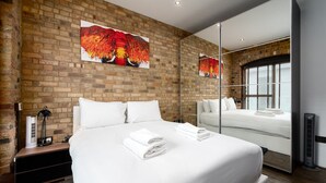 Camera d'albergo con parete in mattoni, un letto grande con lenzuola bianche, un comodino con una lampada e un ampio specchio che riflette l'ambiente.