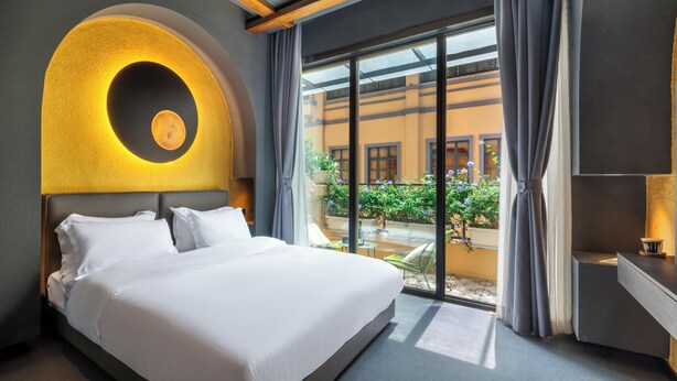 A hotel room with a large bed, a circular light fixture above the bed, a view of a courtyard with plants and a building, and a small table with a cup. - AM Manye Hotel (Kunming)
