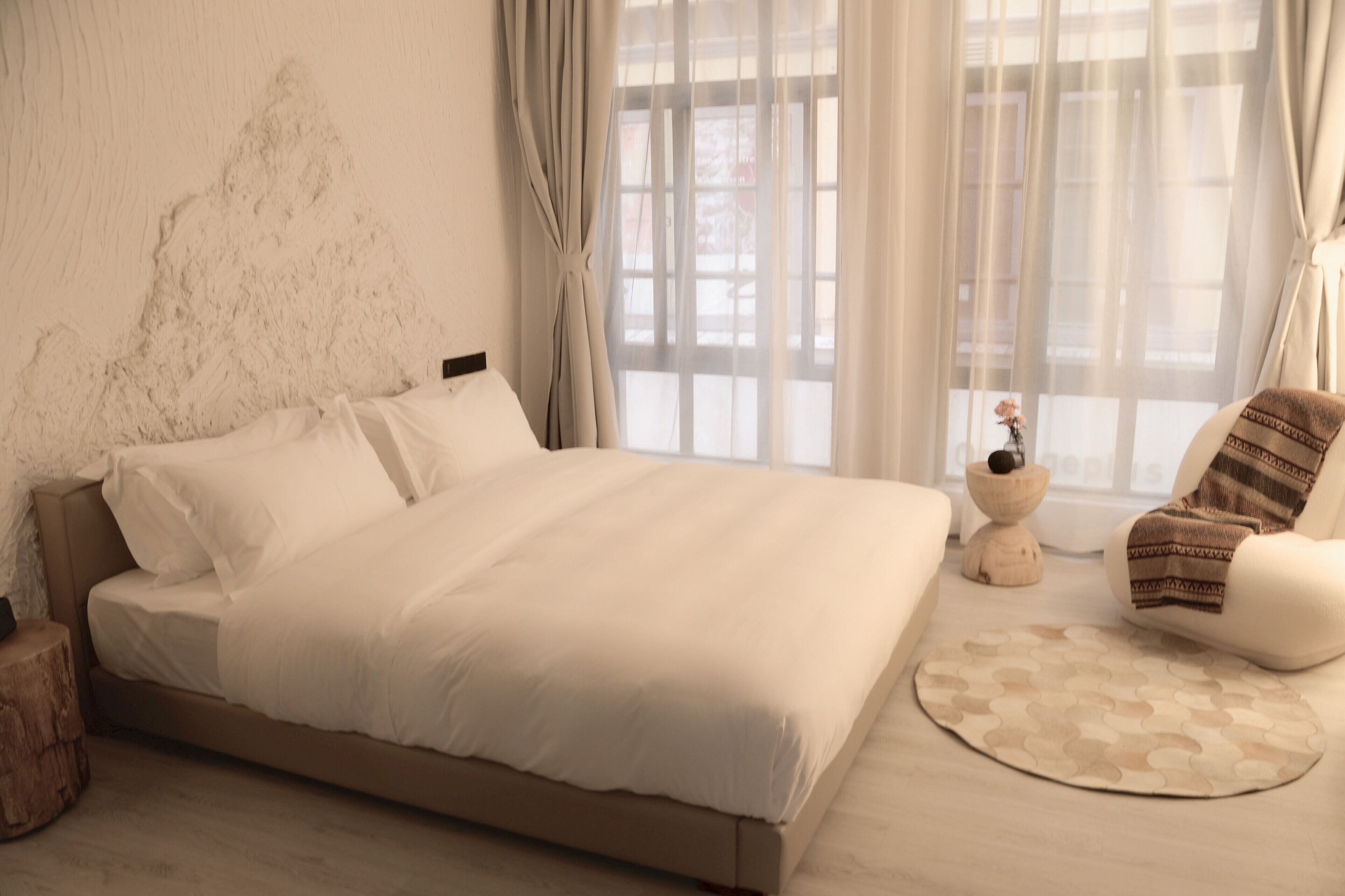 A bedroom with a large bed, a wooden nightstand, a round rug, a chair, and a window with sheer curtains.
