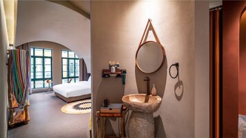 Un dormitorio con cama, espejo redondo, lavabo de piedra y un estante pequeño con artículos de tocador.