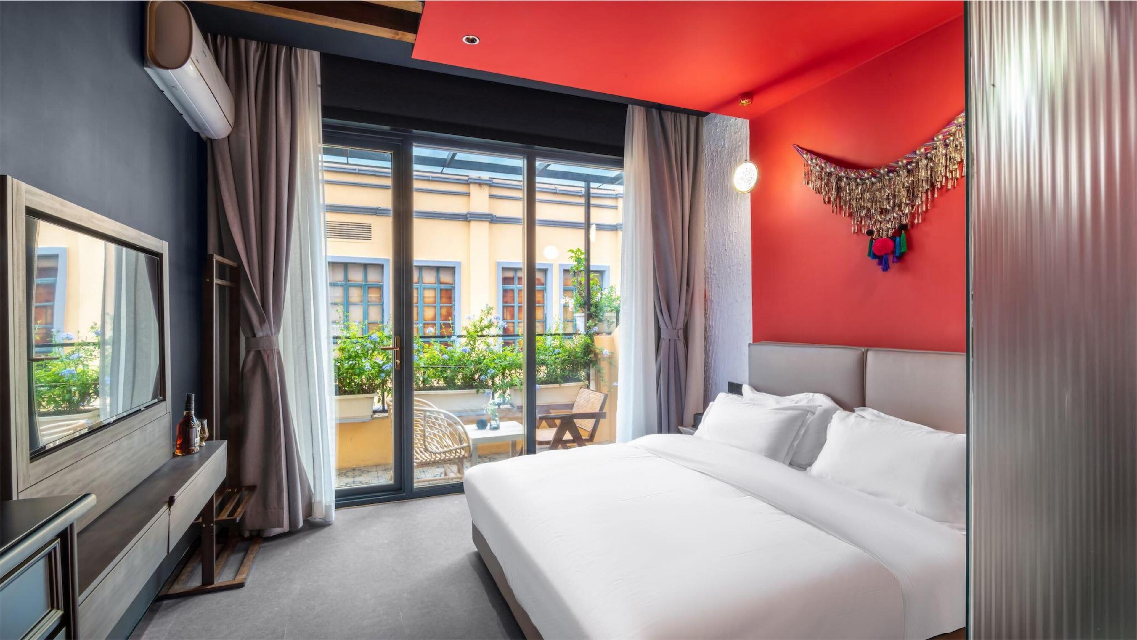 Quarto de hotel com uma cama grande, televisão e varanda com vista para edifícios e vegetação.