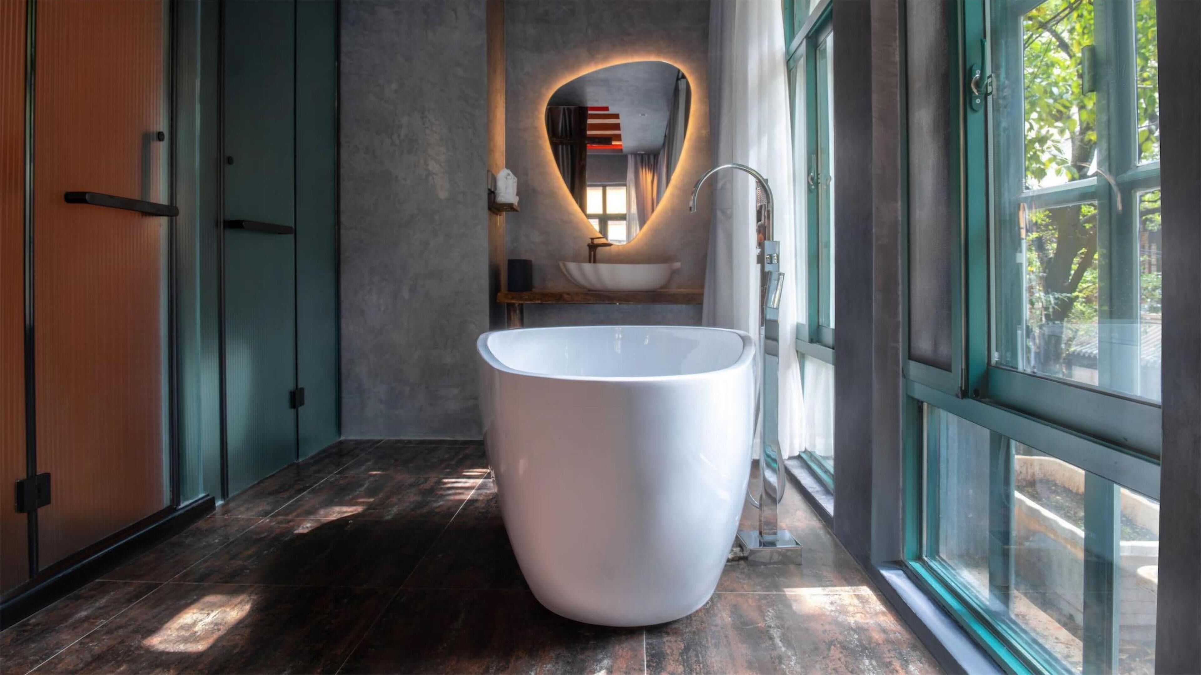 Un baño moderno con una bañera blanca grande, un espejo con luz en forma de corazón y vistas al verdor exterior.