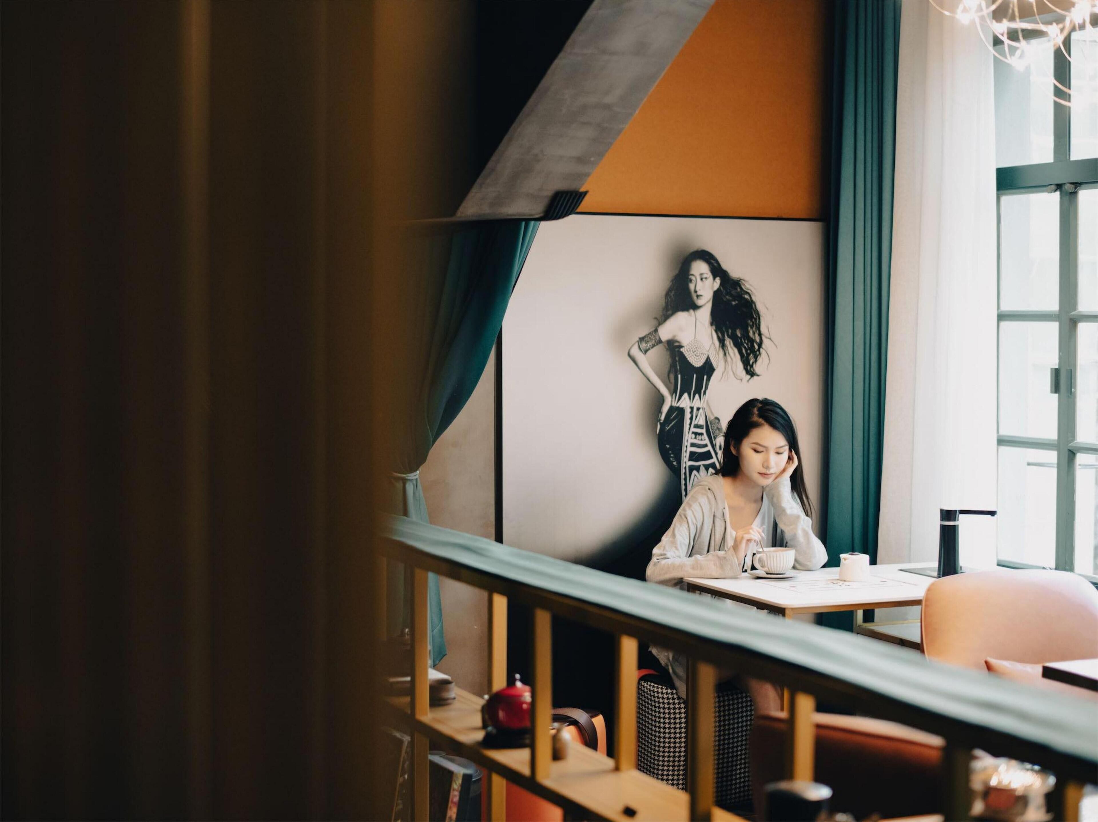 A person sitting at a table in a restaurant with a modern interior, including a large window, a wall with a black and white artwork, and a staircase with a railing.
