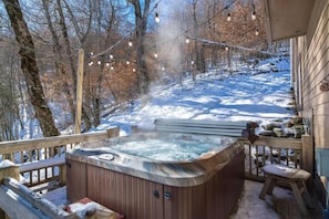 A hot tub surrounded by a wooden deck, with a snowy landscape and trees in the background.