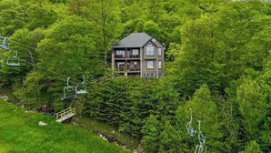 A two-story house with a balcony, surrounded by lush greenery and a ski lift in the background. - Large, Ski-in/Ski-Out, Hot Tub, Wet Bar, Sleeps 16 (Beech Mountain)