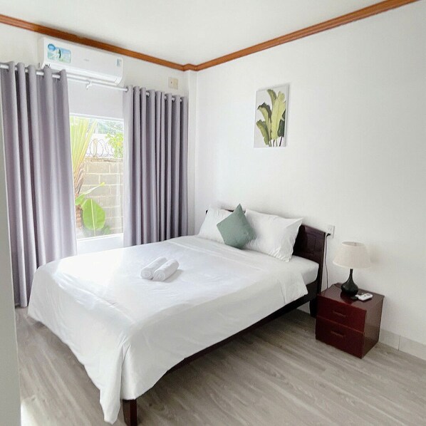 A neatly made bed with white linens and a green pillow, a bedside table with a lamp, and a window with curtains. - Inner Peace Homestay Phan Thiet  (Mui Ne)