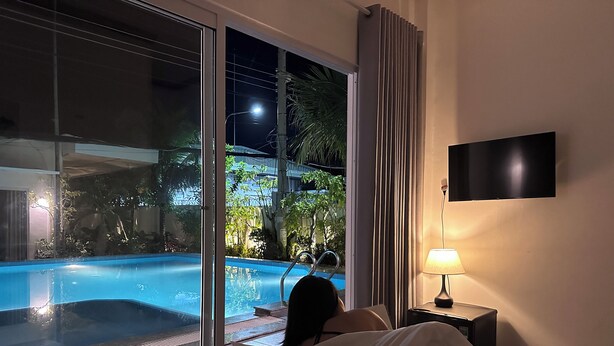 A bedroom with a large window overlooking a swimming pool, a bedside lamp, and a flat-screen TV on the wall. - Inner Peace Homestay Phan Thiet  (Mui Ne)