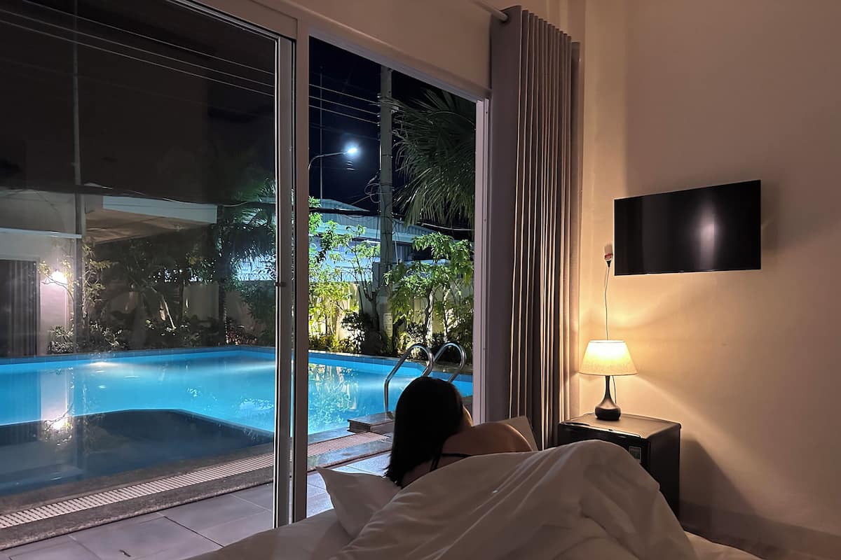 Ein Schlafzimmer mit großem Fenster und Blick auf einen Swimmingpool, einer Nachttischlampe und einem flachbildschirmfernseher an der Wand.