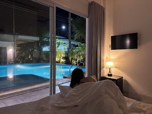 A bedroom with a large window overlooking a swimming pool, a bedside lamp, and a flat-screen TV on the wall. - Inner Peace Homestay Phan Thiet  (Mui Ne)