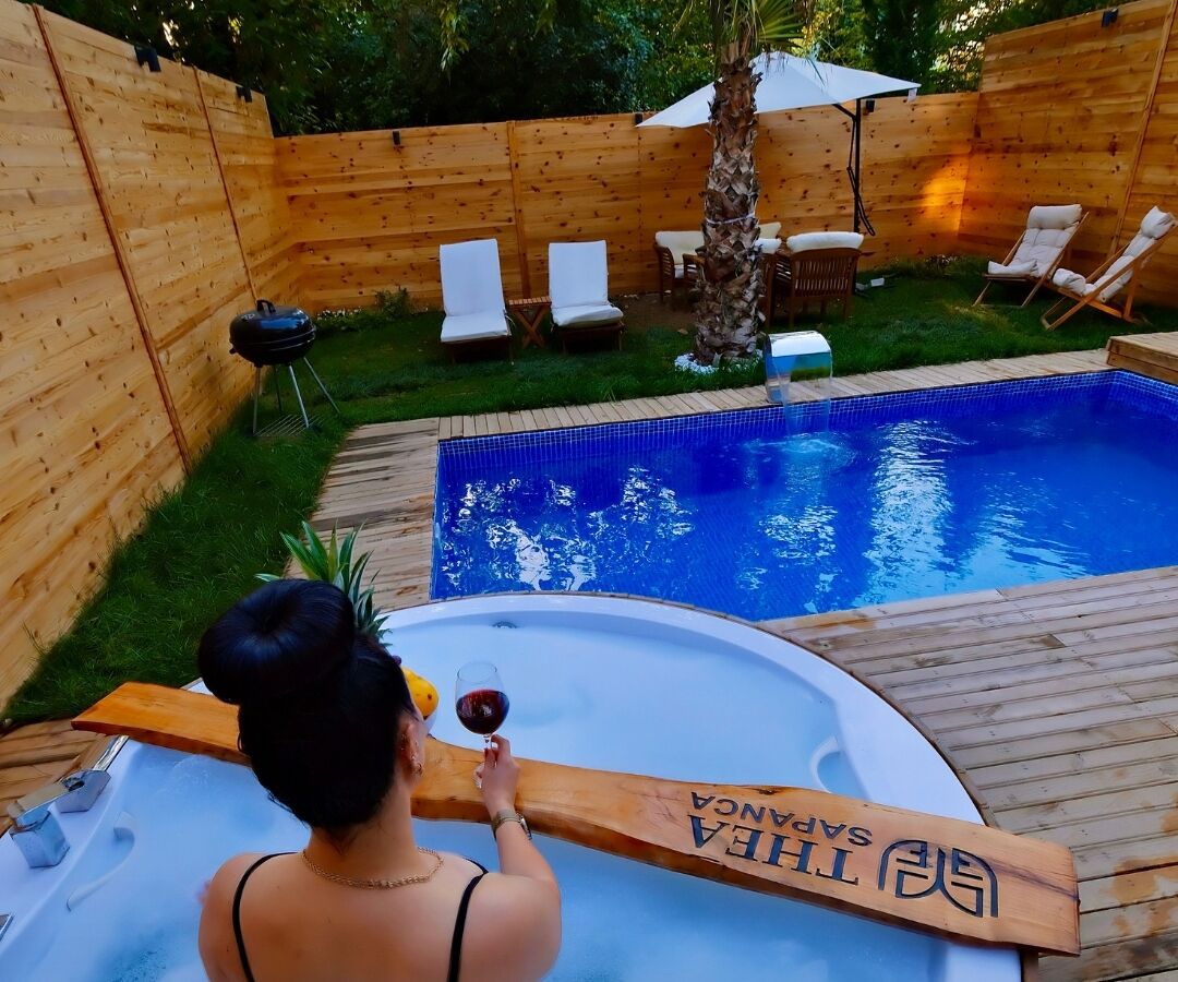Un bain à remous avec une terrasse en bois, entouré d’une clôture en bois, et une piscine avec des chaises longues.