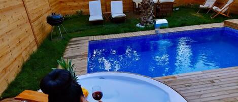 Un bain à remous avec une terrasse en bois, entouré d’une clôture en bois, et une piscine avec des chaises longues.