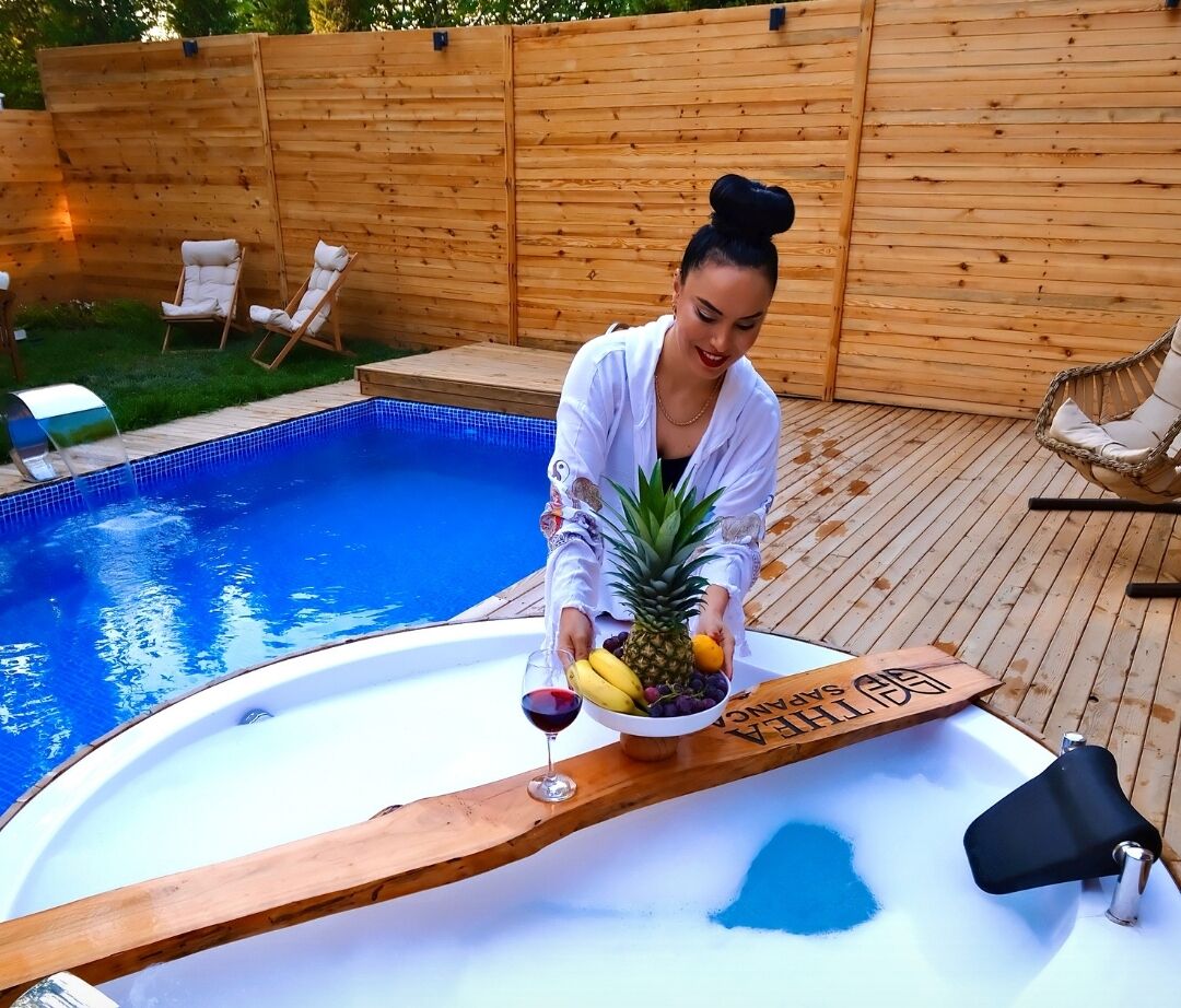 Una persona de pie junto a una piscina con cubierta de madera, sosteniendo un cuenco de fruta y una copa de vino tinto.