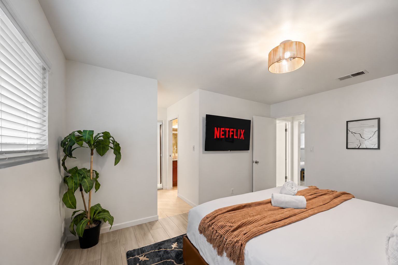 A modern bedroom with a bed, a potted plant, a television displaying 'NETFLIX', and a door leading to another room.