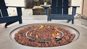 Ein Feuerkorb aus Beton mit einem Metallring und brennendem Holz, umgeben von einer Terrasse mit zwei blauen Adirondack-Stühlen und einem Steintisch.