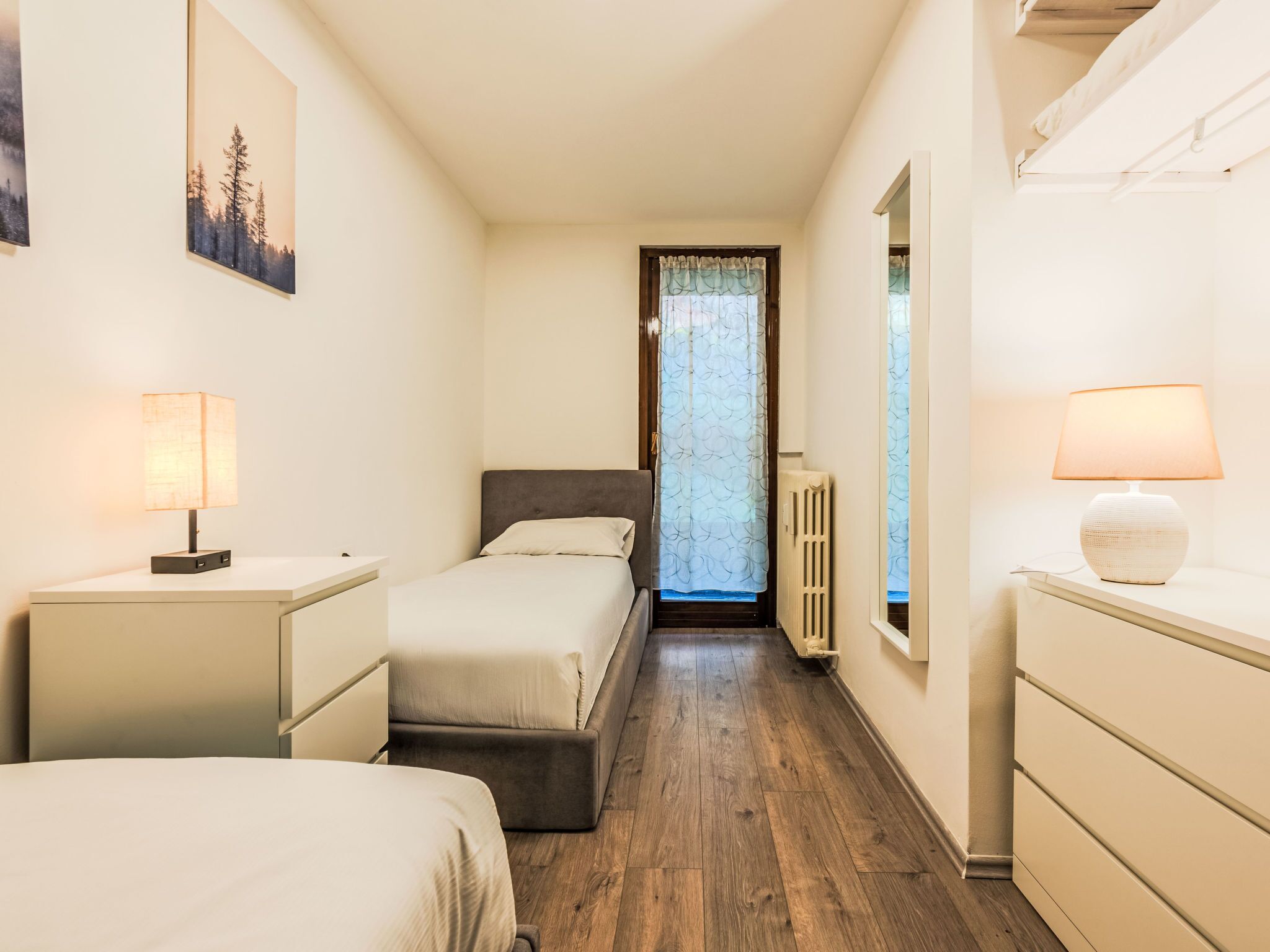 Habitación de hotel estrecha con dos camas, un radiador y una ventana con cortinas.