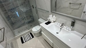 Et moderne badeværelse med et glasvægshus, et hvidt håndvask med et spejl over, og et toilet med en stribet måtte foran.