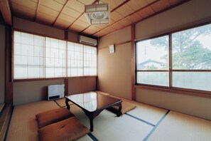 A traditional Japanese room with tatami flooring, a low table, cushions, and large windows with a view of a house and trees.