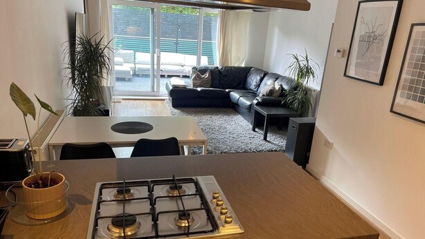 A modern kitchen with a gas stove, a dining table, and a black leather sofa in the background. - 1Bedroom - Sleeps 2 - Terrace, Bbq, Near Transport (London)