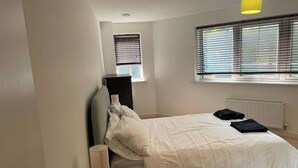 A bedroom with a bed, a bedside table, a lamp, a radiator, and a window with blinds. - 1Bedroom - Sleeps 2 - Terrace, Bbq, Near Transport (London)