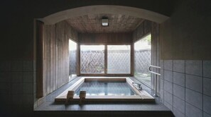 Ein traditionelles japanisches Onsen mit Holzinnenraum, einem Steine-Thermalbad und Blick auf einen von einer Mauer umgebenen Garten.