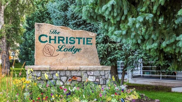 A stone sign with "The Christie Lodge" written on it, surrounded by a garden with various colorful flowers and a large evergreen tree in the background. - Christie Lodge Winter's Best. (Avon)