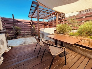 En träaltan med ett bord och två fåtöljer i rotting, en pergola och en trädgårdsdel med växter och en mur av tegelsten.