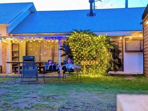 Une maison dotée d’un espace extérieur aménagé avec un coin salon couvert, d’un petit arbre décoré de guirlandes lumineuses et d’une enseigne sur laquelle on peut lire : « DOG LEG ».