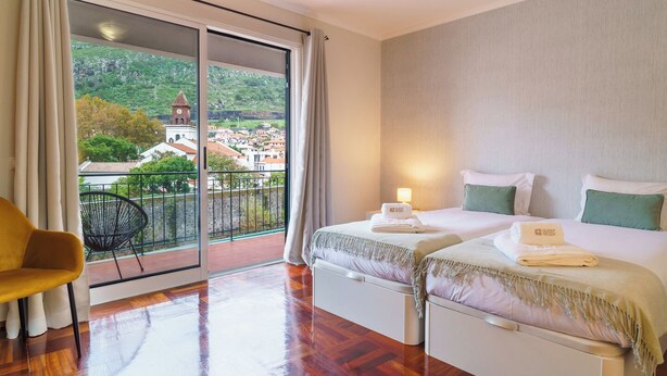 Ein Hotelzimmer mit Balkon, zwei Betten, einem Sessel und Blick auf eine Stadt und eine Kirche.