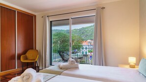 Ein Hotelzimmer mit zwei Betten, einem gelben Sessel, einem Balkon mit Blick auf Grünflächen und Gebäude sowie einer Lampe auf dem Nachttisch.