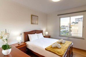 A neatly arranged hotel room with a bed, bedside tables, a window with a city view, and a potted plant. - GuestReady - Charming retreat in Funchal (Funchal)