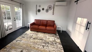 A room with a brown leather sofa, a patterned rug, a white wall with decorative wall art, and a large window with curtains. - Charming backyard Guesthouse with 2 bedrooms and WiFi, AC in Pleasant Hill (Pleasant Hill)