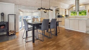 Una cocina moderna con una isla central, un comedor con cuatro sillas, un hogar a leña y un amplio ventanal con vistas al verdor.