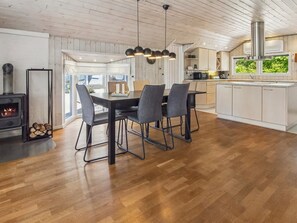 A modern kitchen with a central island, a dining area with four chairs, a wood-burning stove, and a large window with a view of greenery.