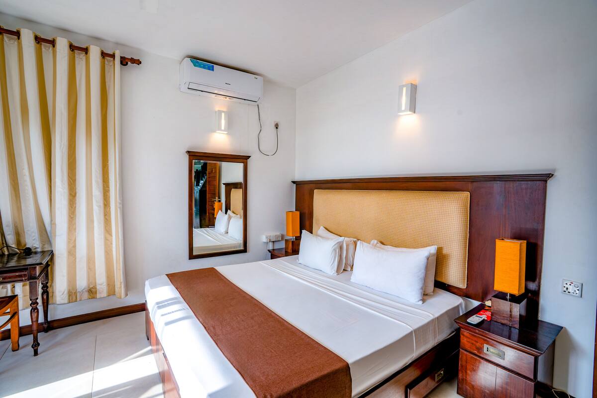 A hotel room with a large bed, a wooden headboard, a nightstand with a lamp, a desk with a chair, a mirror, and an air conditioning unit.