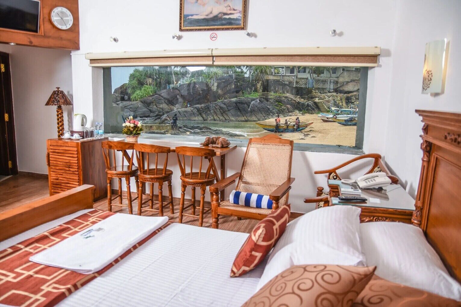 Una habitación de hotel con cama, escritorio con sillas, ventana con vista a una playa rocosa y barcos, y teléfono sobre el escritorio.