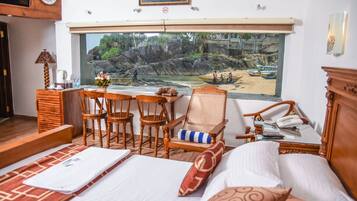 Una habitación de hotel con cama, escritorio con sillas, ventana con vista a una playa rocosa y barcos, y teléfono sobre el escritorio.