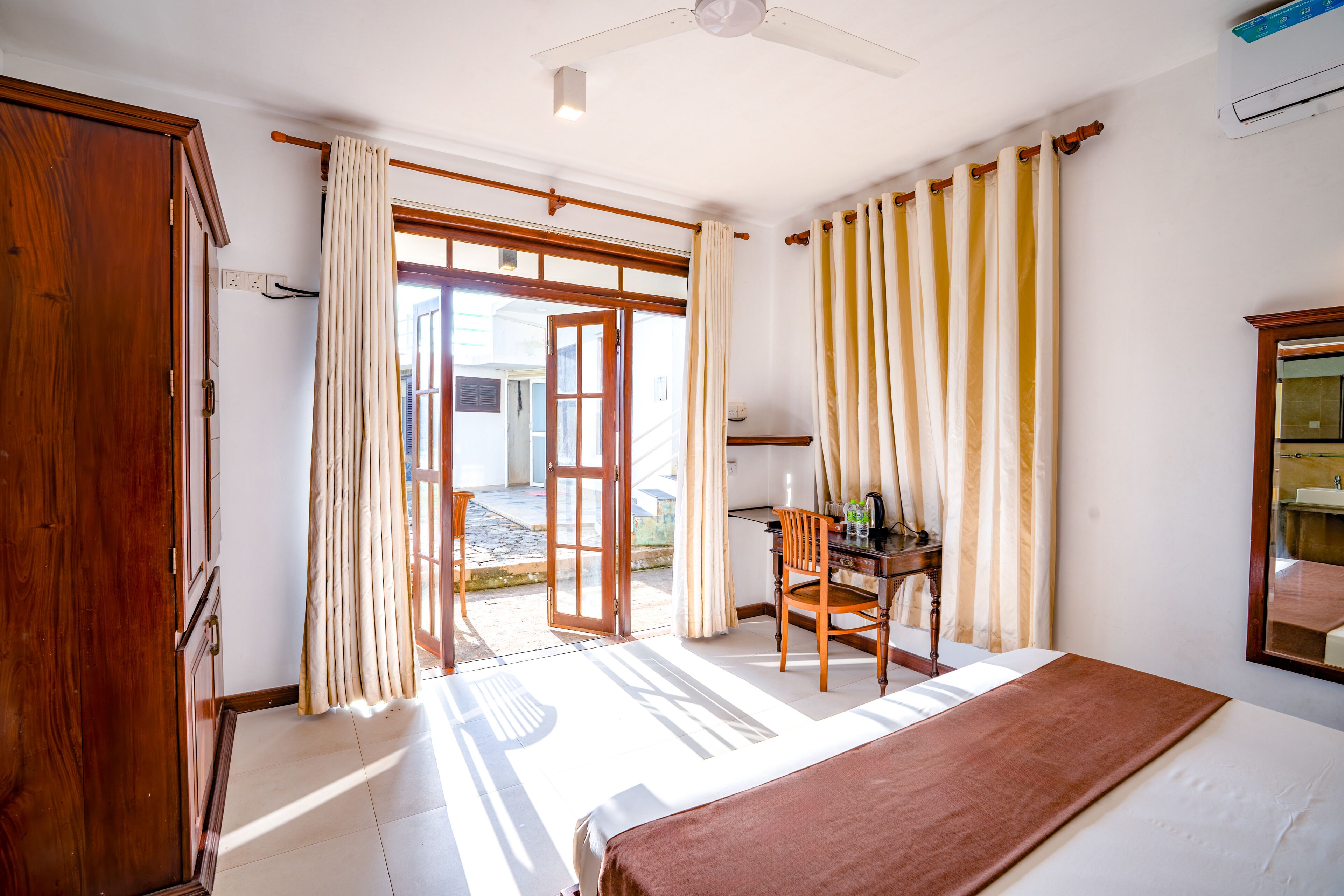 Habitación de hotel con cama, escritorio y silla. Cuenta con una ventana con cortinas, una puerta que da al exterior y un ventilador de techo.